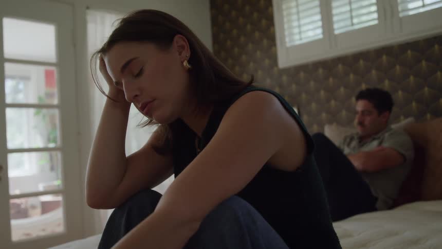 Woman sitting on the edge of the bed with a weary expression while a man stays in the background