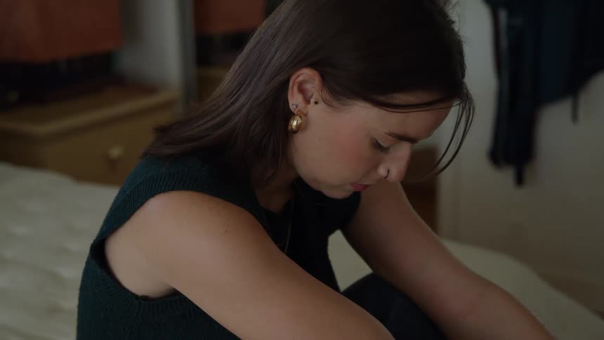 Young woman sitting down on a bad looking down with a sad face expression indoors during day time