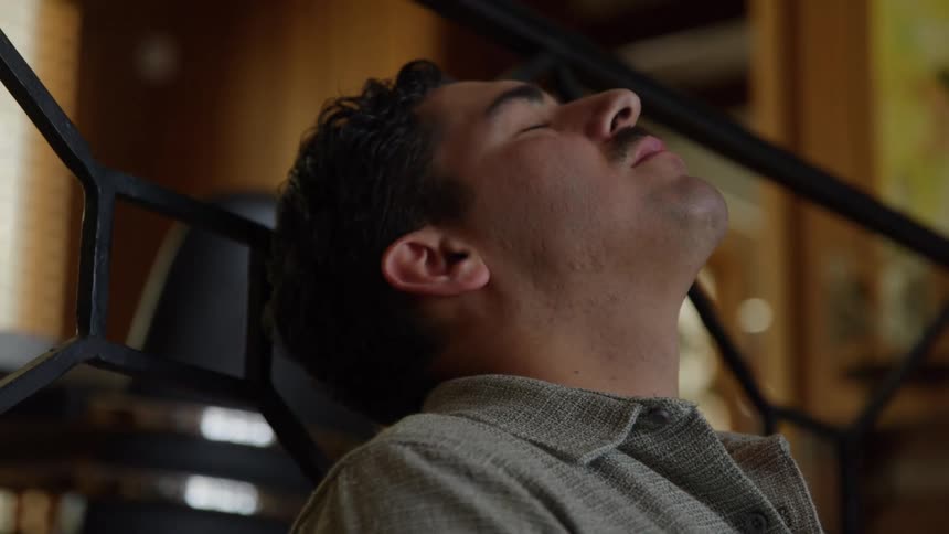 A close view shows a man with dark curly hair and a mustache wearing a gray collared shirt relaxing with his eyes closed and his head tilted back in a bright wood paneled room