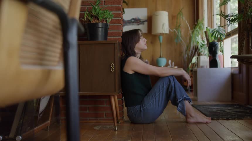 Woman sitting on the floor leans against the wall and gazes out the window in silence
