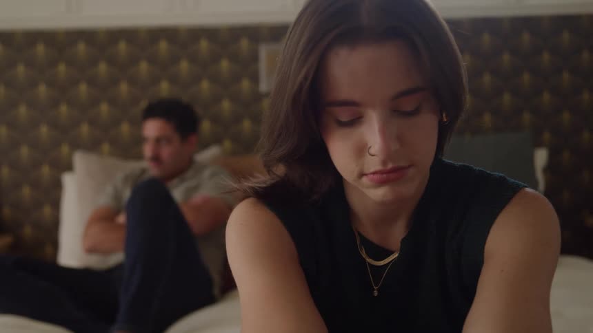 The video is a closeup of a young woman with long dark hair sitting on a bed looking down with a troubled or pensive expression in the blurred background a man is visible sitting further back on the bed with his back partially turned suggesting tens