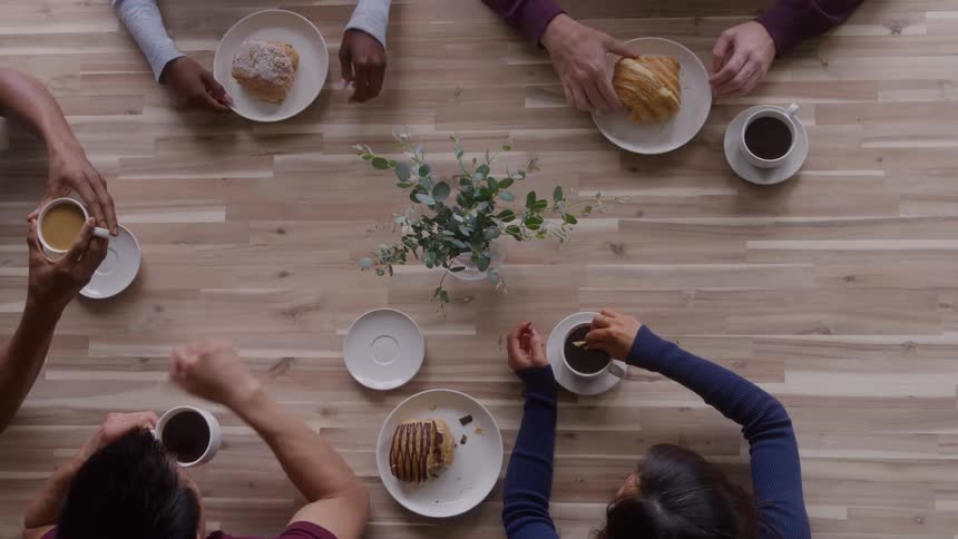 The video is an overhead shot of a group of people sitting at a wooden table enjoying coffee and pastries the hands visible on the table suggest a casual gathering or business meeting during a light break
