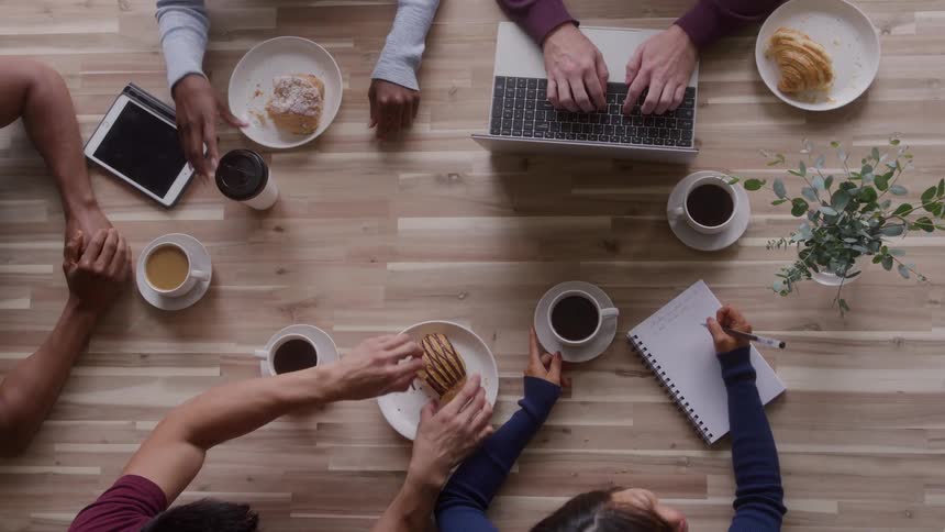 Various hands share coffee pastries and technology over a wooden table