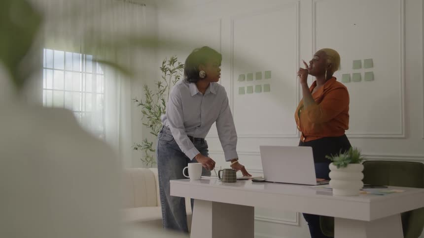 Two women talk in a modern office with natural light and sticky notes on the wall