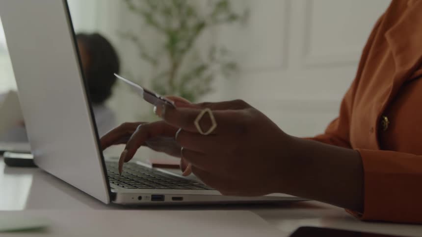 Hands of a black woman wearing an orange suit typing on a laptop computer while holding a card on her other hand with another black woman sitting on the background indoors during day time