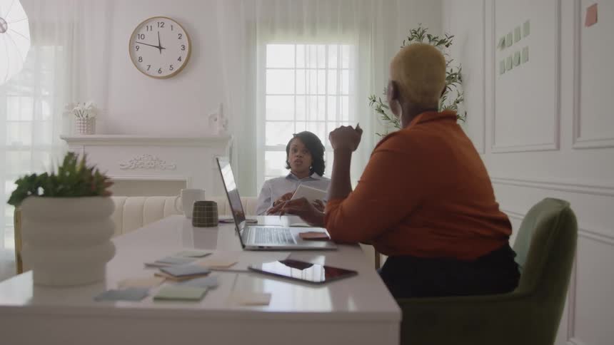 Two women sit across a white table from each other in a brightly lit room the woman in the foreground faces away and gestures while speaking one woman holds a tablet and looks at the speaker a laptop and various office supplies are on the table