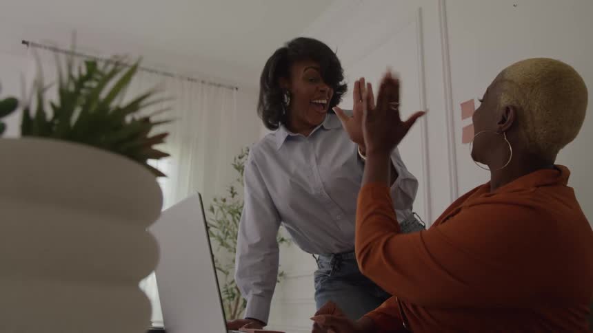 Two young black women celebrating while working together with a laptop at a desk in an office during daytime