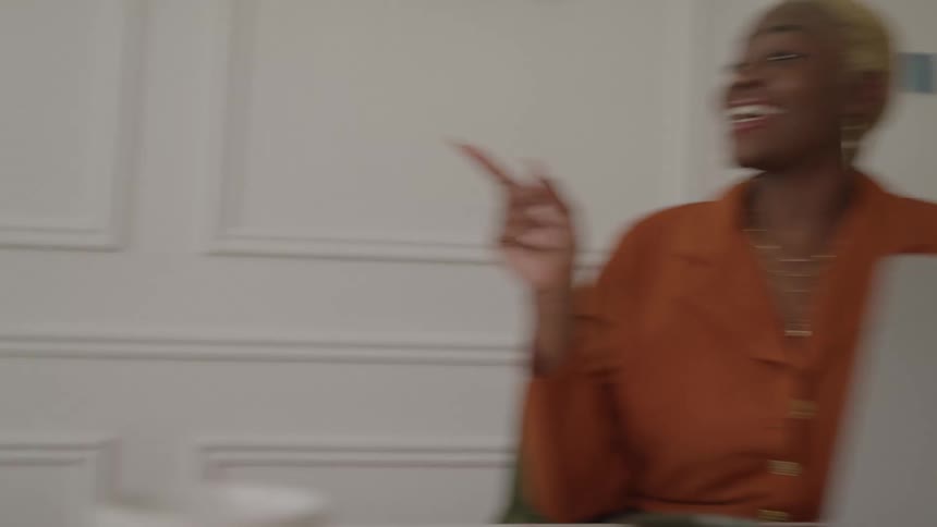 A camera pans between two women engaged in a work discussion the first woman sits on a couch holding a tablet and gesturing while speaking the second woman sits at a desk with a laptop and smiles while listening