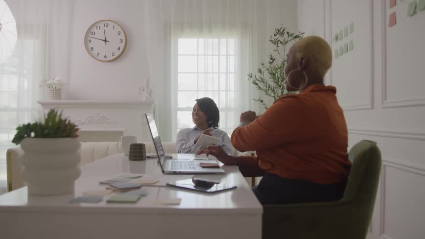 Two women work together in a bright office discussing ideas in front of their laptops