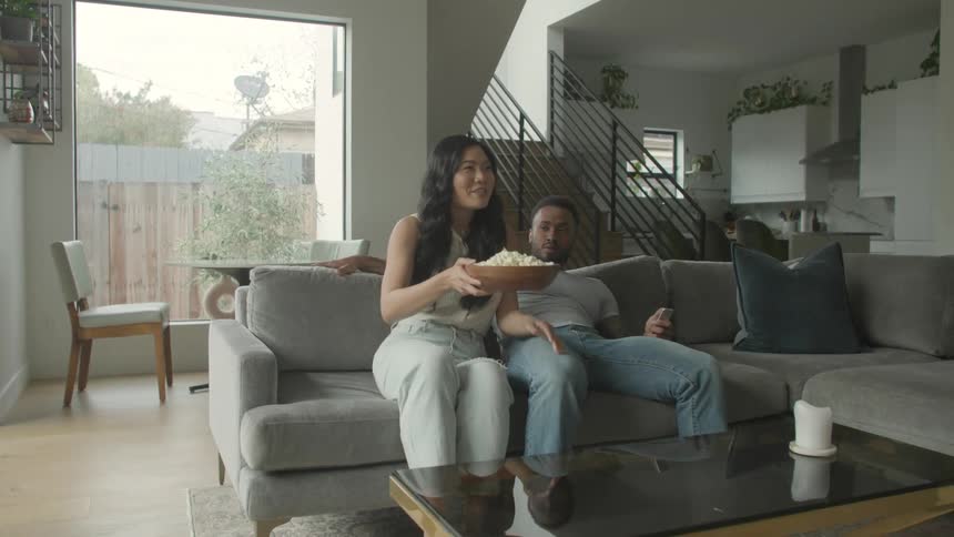 Young black man and young biracial woman sitting down on a couch while eating popcorn and hugging indoors during day time