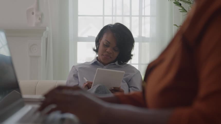 A person typing on a laptop keyboard in the foreground another woman in the background sits on a white couch and writes on a digital tablet the room is brightly lit by a large window a tall indoor plant stands on the right