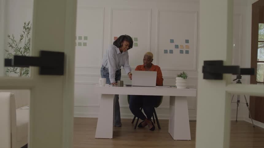 A view through an open doorway shows two women working at a white desk one woman stands and leans over a laptop the other woman sits and looks at the standing woman the background wall is decorated with small colorful squares