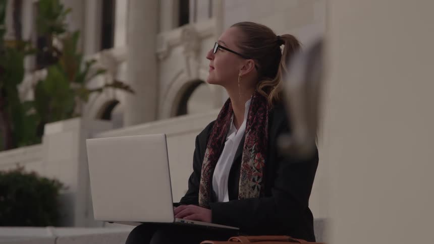 Woman sitting outside typing on a laptop computer on her knees as she stops to look around