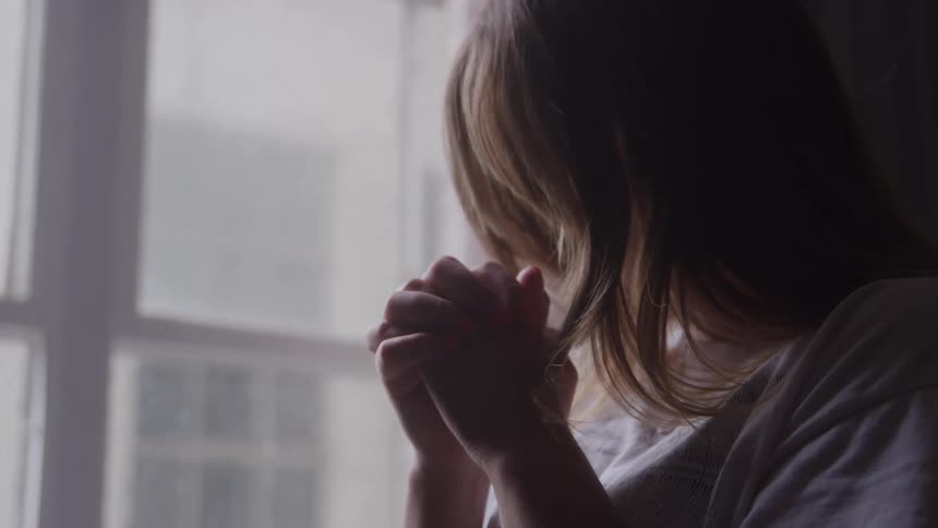 A young woman stands beside a window with her hands clasped together in prayer at midday