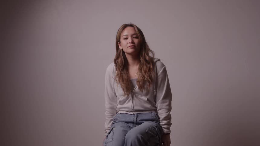 The video features a mediumclose shot of a young woman entering the frame and sitting down on a small stoolshe is wearing a light gray hooded sweatshirt and cargo jeans and she sits looking directly at the camera with a neutral and composed expressio