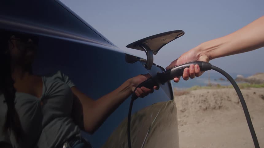 Woman plugs an electric car charger in to an automobile parked on the coast