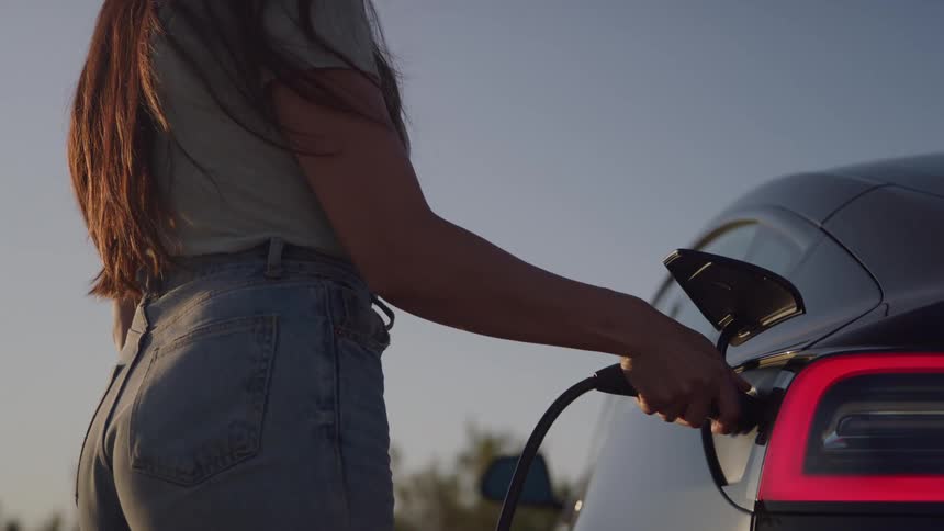 A woman wearing a white shirt and jeans plugs a charging cable into the rear port of a dark electric car with a blurry wind turbine spinning in the sunset background