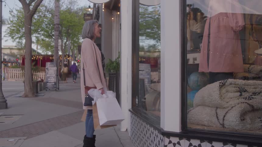 The video shows an attractive middleaged woman with silver hair happily walking down a sidewalk while carrying several shopping bagsshe briefly pauses to look at the clothing and home goods display in a large storefront window before continuing to wa