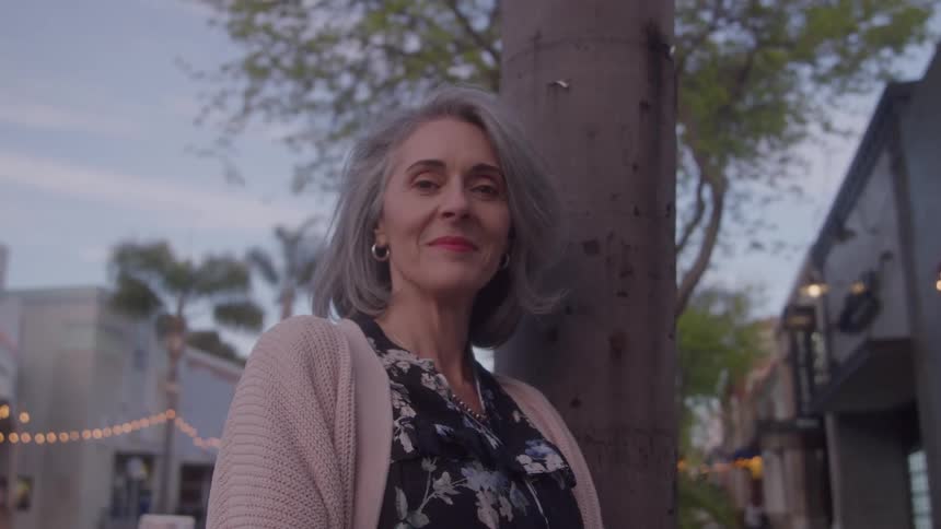 A woman with gray hair and earrings stands near a pole she looks forward and smiles the background shows a street with buildings lights are visible on the street all bathed in natural sunlight setting with a cloudy sky