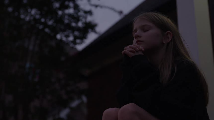 A young girl with long straight lightcolored hair is sitting outdoors in the dim light of dusk or dawn appearing pensive or distressed as she holds her hands to her chin she is positioned on a porch or step against a dark shadowy background of trees