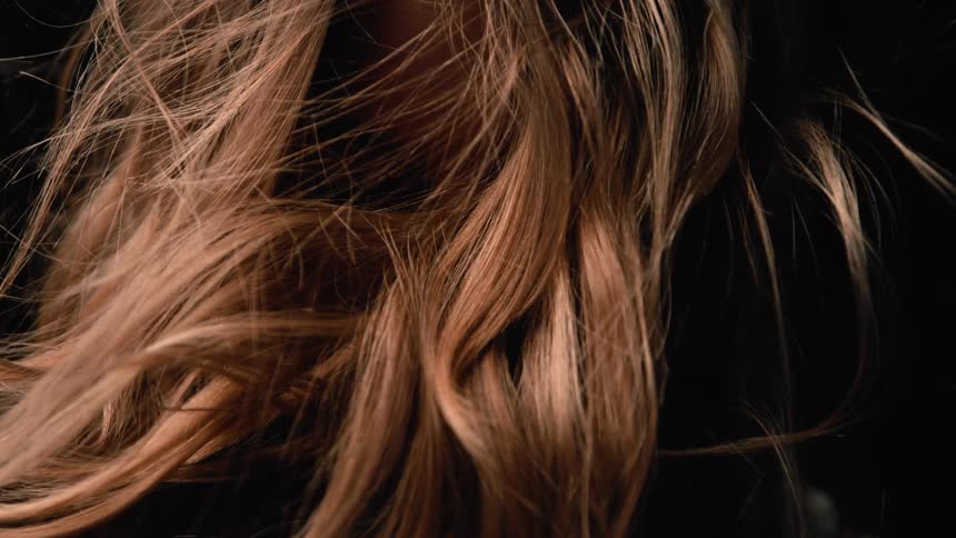A closeup shot focuses on a mass of long brownishblonde hair showing a mix of loose waves and straight strands the hair is set against an entirely dark background highlighting its texture and color as it is softly illuminated and slightly ruffled