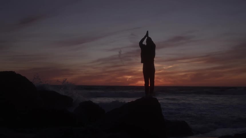 A person stands on a rock holds hands above his head in a pray position the sun sets over the ocean waves move toward the shore the man is silhouetted against the light