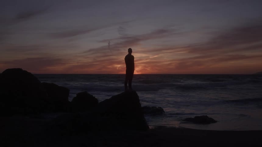 A man stands on a rocky shoreline watching the waves crash against the rocks as the sky glows orange and gradually darkens at sunset