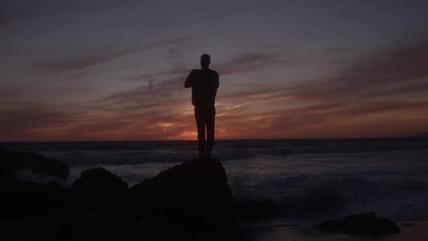 A man stands on a rock by the shoreline at sunset with the sky glowing orange as it gradually fades into darkness