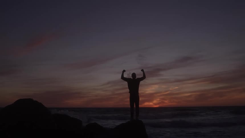A man stands on a beach with his hands raised as the orange sunset sky glows behind him