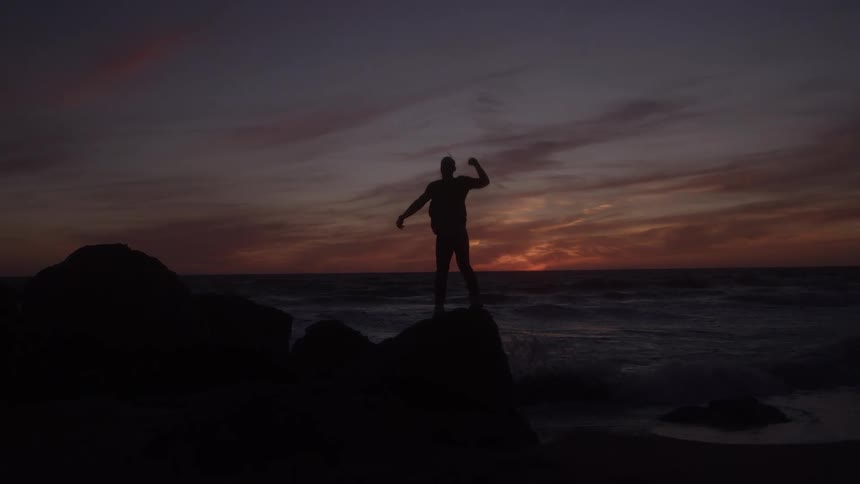 The video features a dramatic silhouette of a person standing atop large rocks on a beach at sunsetthe person raises one arm in a celebratory or victorious pose against the vibrant dark orange and purple hues of the twilight sky over the ocean