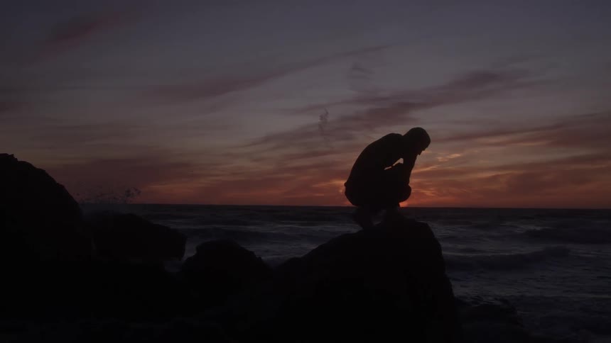 Man bending down a rock along the coast stands as waves roll into shore