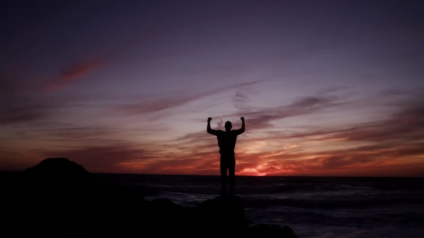 A man stands on a beach with his hands raised as the orange sunset sky glows behind him
