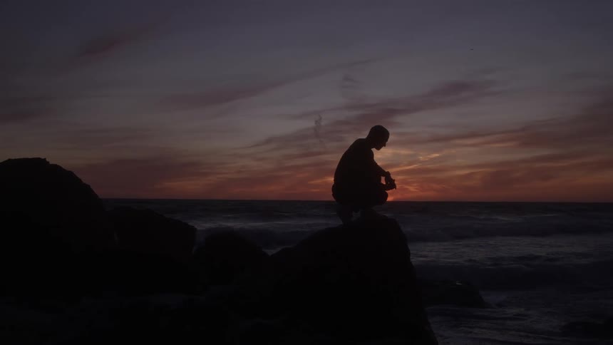 A silhouetted man is crouched down on a dark rocky jetty or breakwater looking out over the ocean waves against a vivid orange and purple sunset sky