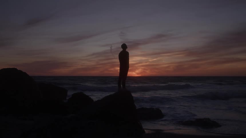 A man stands on a rocky shoreline watching the waves crash against the rocks as the sky glows orange and gradually darkens at sunset