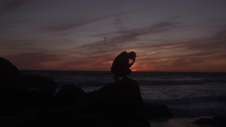 A person crouches on a rock the ocean water moves in waves the sky displays clouds near the horizon the figure is a silhouette against the suns light