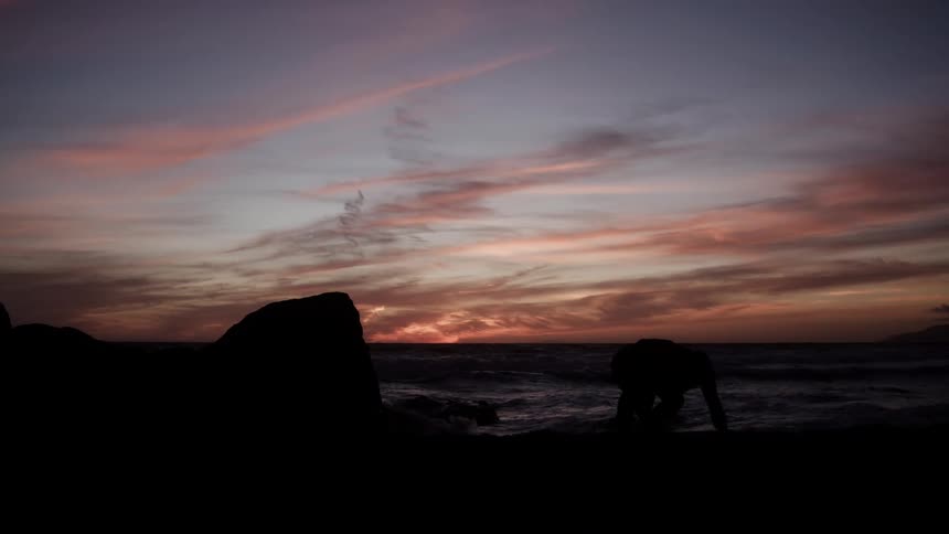Man stands on a rock along the coast and leaps into the air and running across the beach