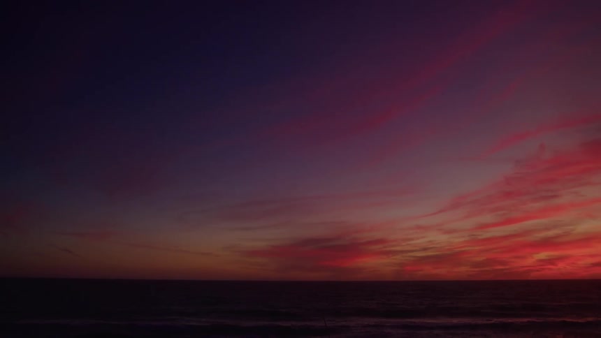 A dark dramatic wide view of the ocean shows intense red and orange clouds fading into a deep purple sky above the horizon line at dusk