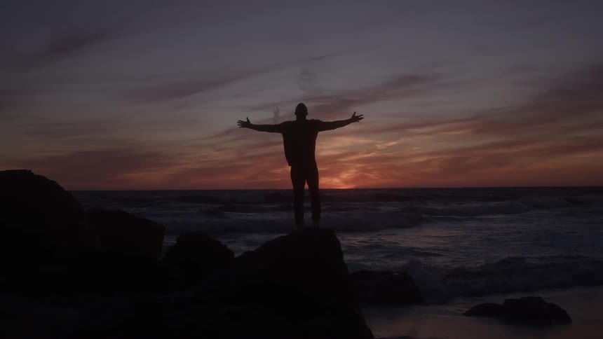 Man standing on a rock along the coast stretches out his arms as waves roll into shore