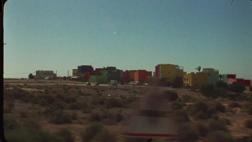 This video shows a wideangle shot of a small community of brightly colored buildings in a dry open landscapethe buildings painted in shades of yellow red blue and green stand out against the clear blue sky and the brown scrubcovered terrain i