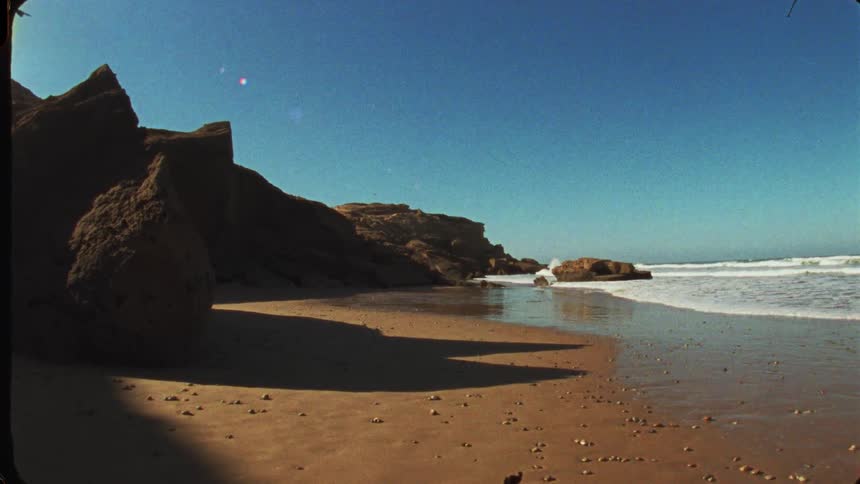 This short video captures a wide shot of a secluded beach cove on a sunny daylarge dark rock formations and cliffs dominate the left side of the frame casting shadows on the wet brown sandon the right the ocean waves are clearly visible crashi