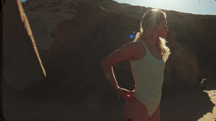 A beautiful woman wearing a light blue one piece swimsuit stands on a sandy beach in bright sunlight posing next to large dark rocks with her hands on her hips