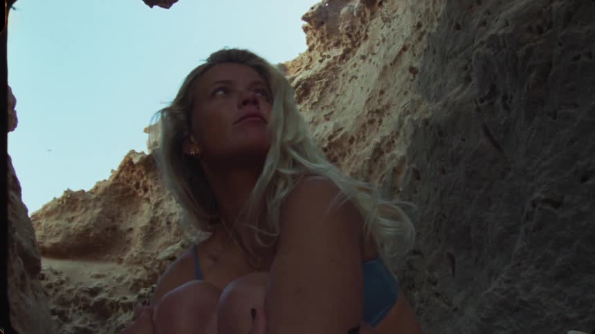 The provided video shows a closeup of a young woman with long blonde hair wearing a bikini top sitting in a narrow passage between natural rock or earth formationsthe lowangle shot and the textured walls create a contrast between the woman and the