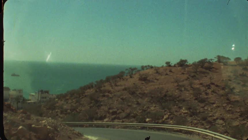 A high angle vintage style view shows a vehicle driving down a winding asphalt road on a dry brown hillside overlooking the bright blue ocean and coastal town below