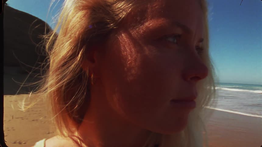 This short video features a closeup profile shot of a young woman with long blonde hair standing on a sunny beachshe is looking out towards the ocean waves with her face framed by the suns glare and her hair gently blowing in the wind
