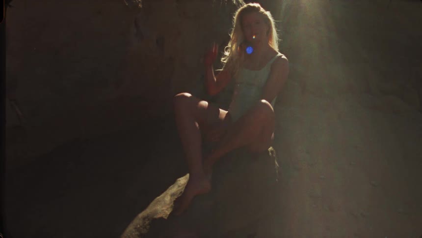 A blonde woman wearing a light blue one piece swimsuit sits on a rock backlit by a bright ray of sunlight shining overhead