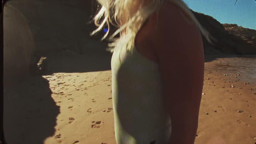 A close view shows a woman with long blonde hair wearing a light green one piece swimsuit standing barefoot on the sandy beach next to a dark rock formation with footprints in the sand
