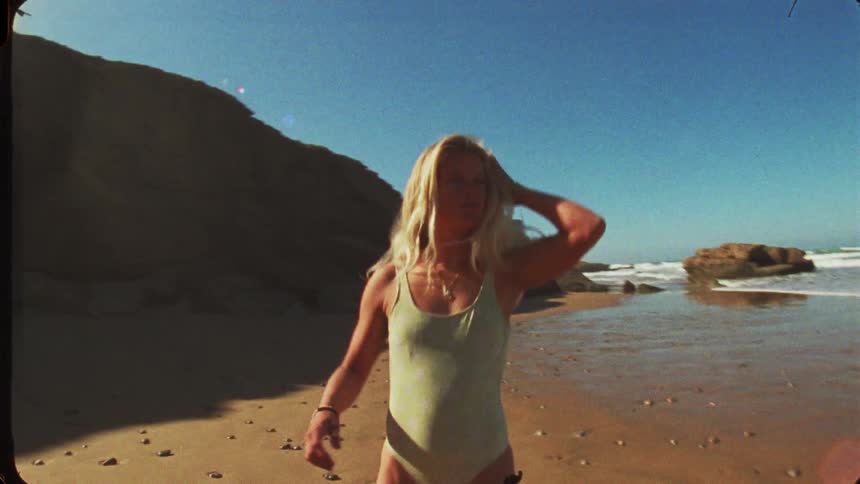 This video follows a young woman with long blonde hair as she walks along a sunny secluded beachshe is wearing a lightcolored lowback onepiece swimsuit and walks in the sand toward the water and a large dark cliff facethe footage has a vinta