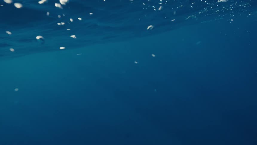This underwater shot features a vibrant deep blue backdrop with a constant stream of bubbles descending from the bright water surface abovethe white illuminated air bubbles drift downward and then dissipate into the darker depths suggesting a contin