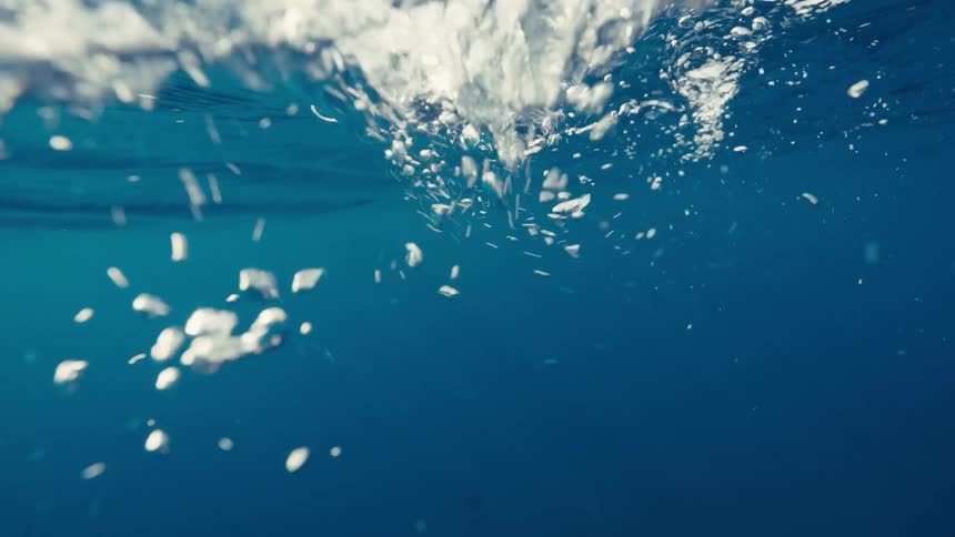 This is an underwater shot in deep blue water focusing on the surface where sunlight penetrates numerous white air bubbles are seen floating and rising toward the top suggesting movement or a disturbance at the waters surface