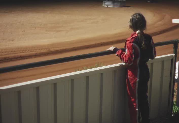 Woman wearing a racing uniform stands watching a go kart race